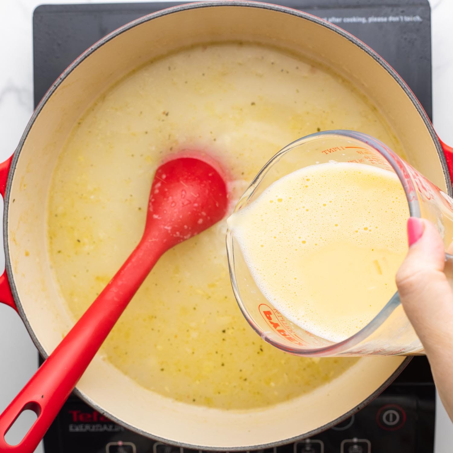 a mixture of eggs and lemon juice added to chicken soup in a dutch oven, stirred with a red spoon.