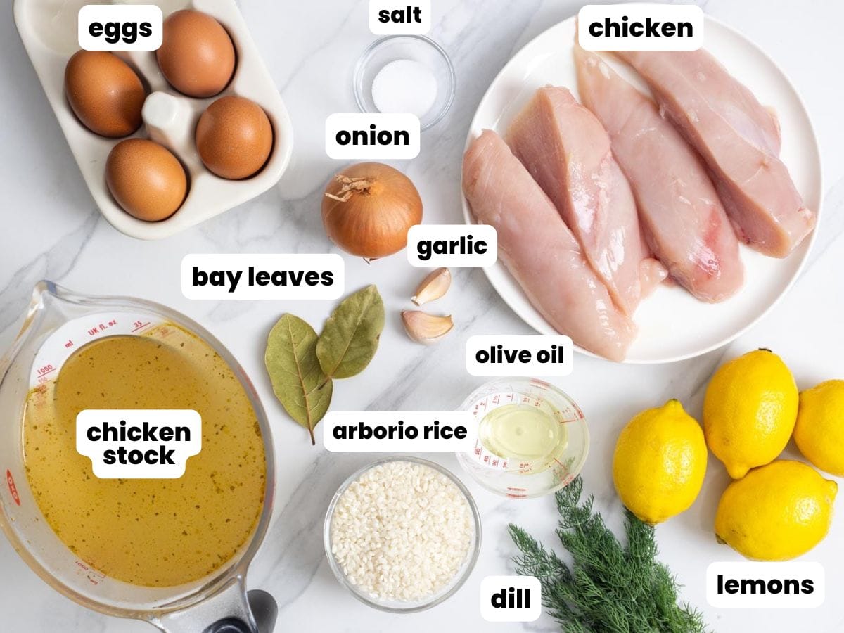 a plate of raw chicken next to four whole lemons, sprigs of fresh dill, arborio rice, olive oil, garlic, onion, eggs, chicken stock, and salt. The ingredients for making avgolemono soup.