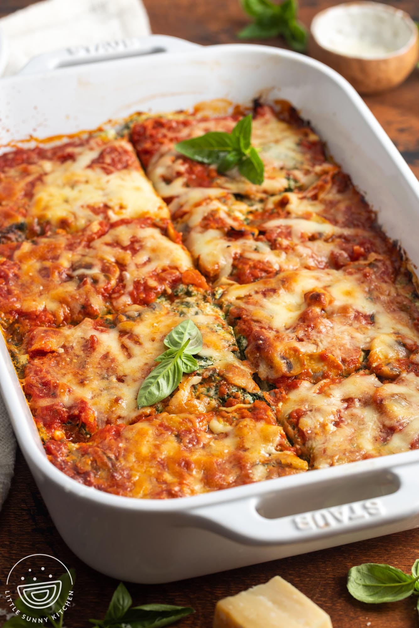 A while ceramic baking dish filled with baked eggplant lasagna, garnished with fresh basil. The lasagna is cut into 8 square servings.