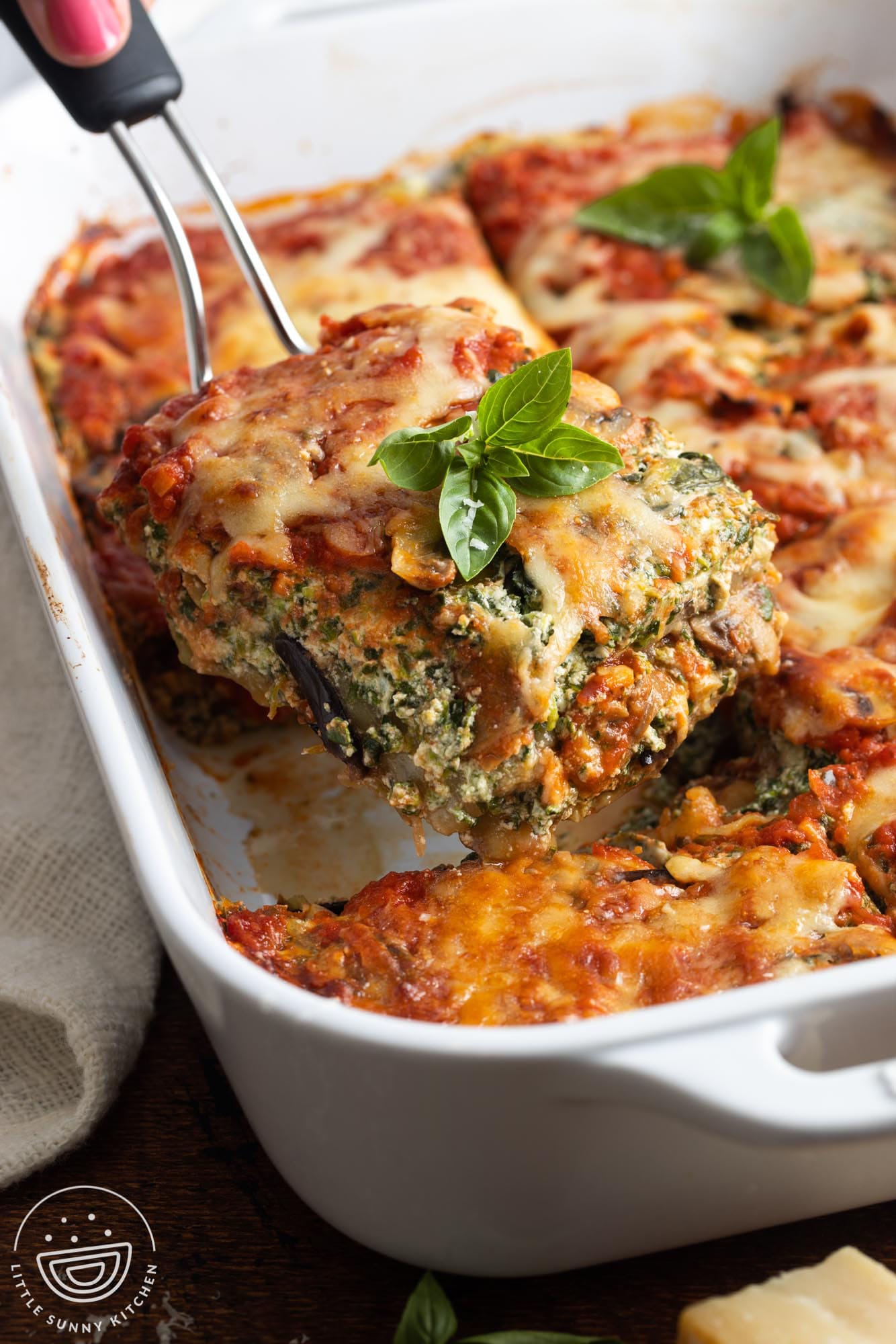 a spatula lifting a square piece of vegetarian eggplant lasagna from a white ceramic baking dish.