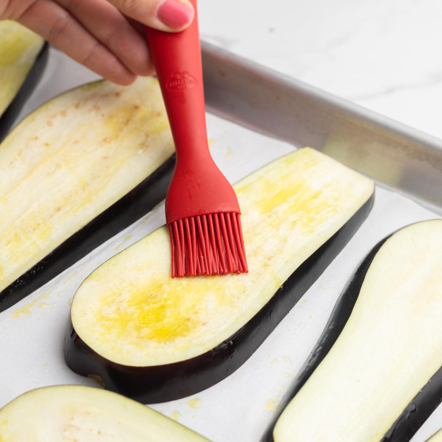 a hand brushing eggplant slices with olive oil using a small red silicone brush.