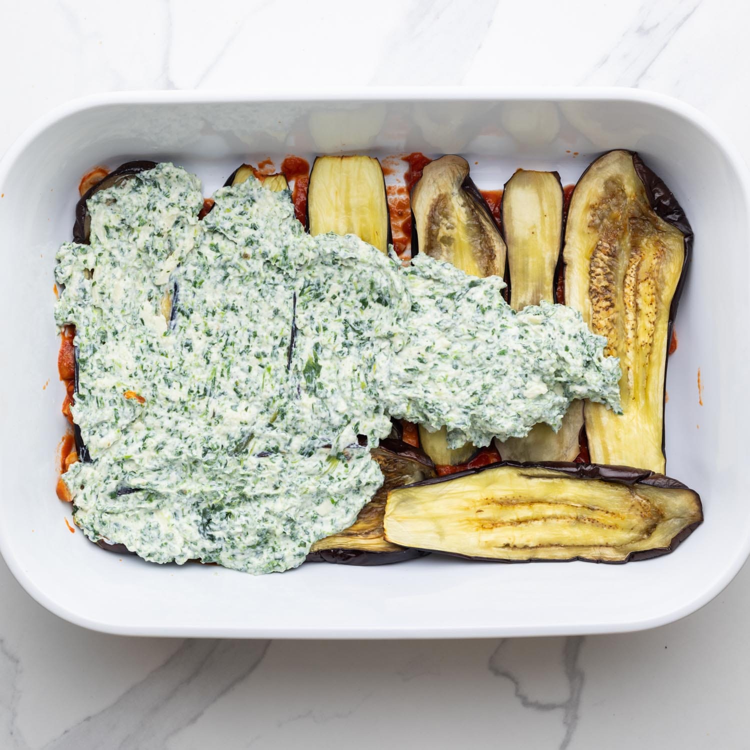 ricotta cheese with herbs spread over roasted eggplant slices in a white baking dish.