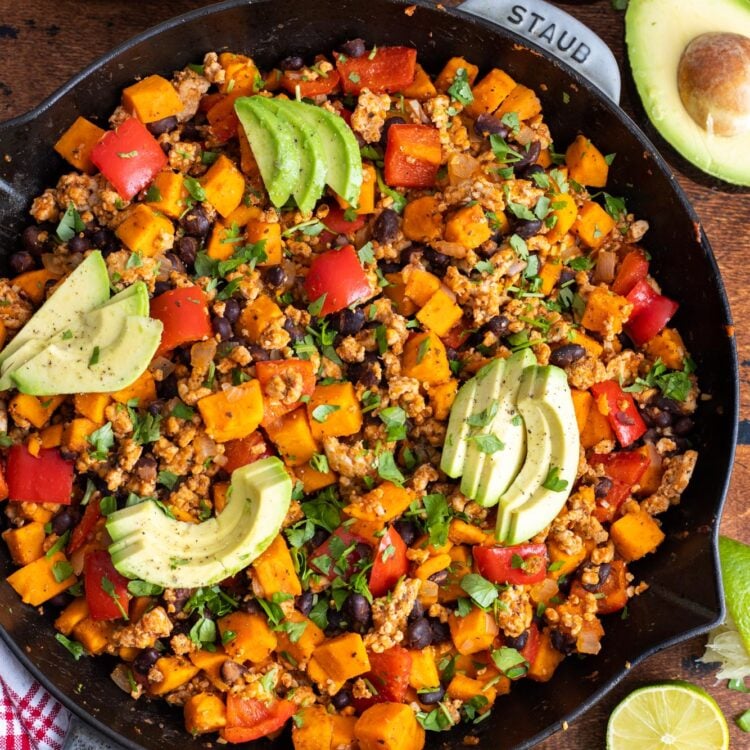 a large cast iron pan filled with ground turkey and sweet potato skillet meal, topped with sliced avocados, viewed from overhead.