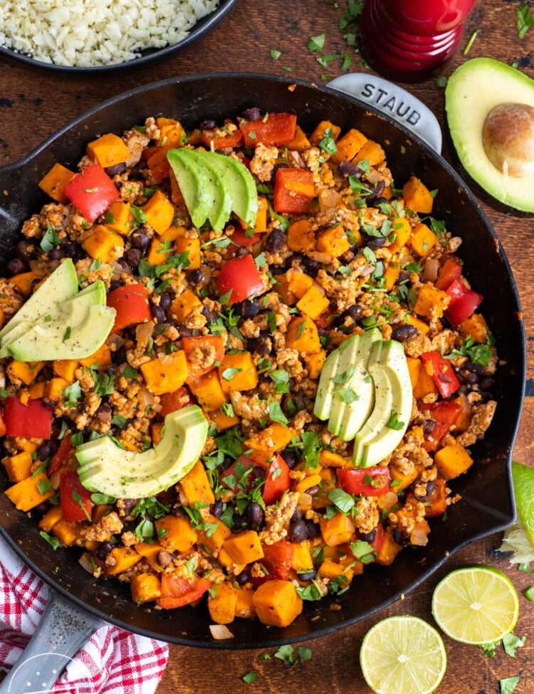 a large cast iron pan filled with ground turkey and sweet potato skillet meal, topped with sliced avocados, viewed from overhead.