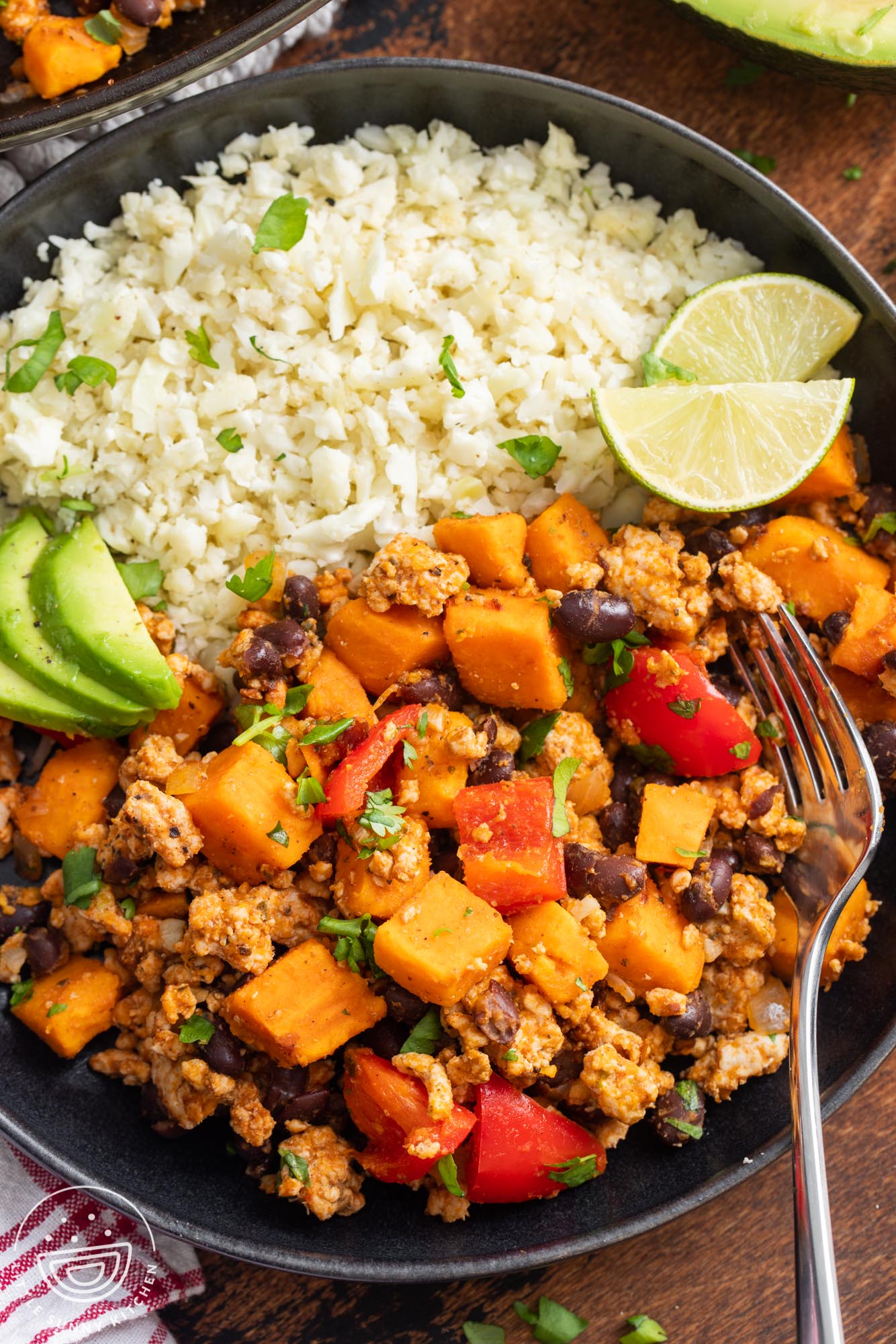 ground turkey with cubed sweet potatoes, black beans, and red peppers, on a plate with cauliflower rice, sliced avocado, and lime wedges.