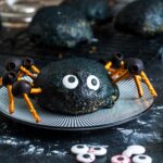 a buffalo chicken bomb which is buffalo chicken and cheese inside a pizza dough ball, decorated black and with pretzel and olive leggs to look like a large spider. The bomb is sitting on a black and white striped plate.