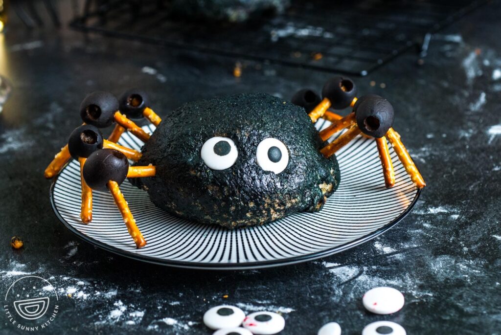 a halloween spider buffalo chicken bomb which is buffalo chicken and cheese inside a pizza dough ball, decorated black and with pretzel and olive leggs to look like a large spider. The bomb is sitting on a black and white striped plate.