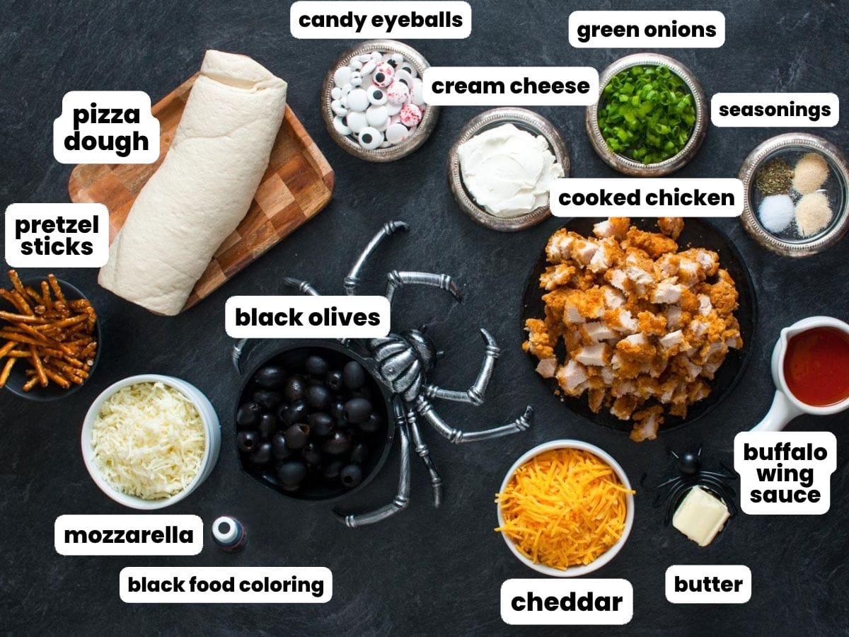 Ingredients for Halloween buffalo chicken bombs in small bowls arranged on a black background. Ingredients include pizza dough, cooked crispy chicken, shredded cheese, black olives, wing sauce, and seasonings.