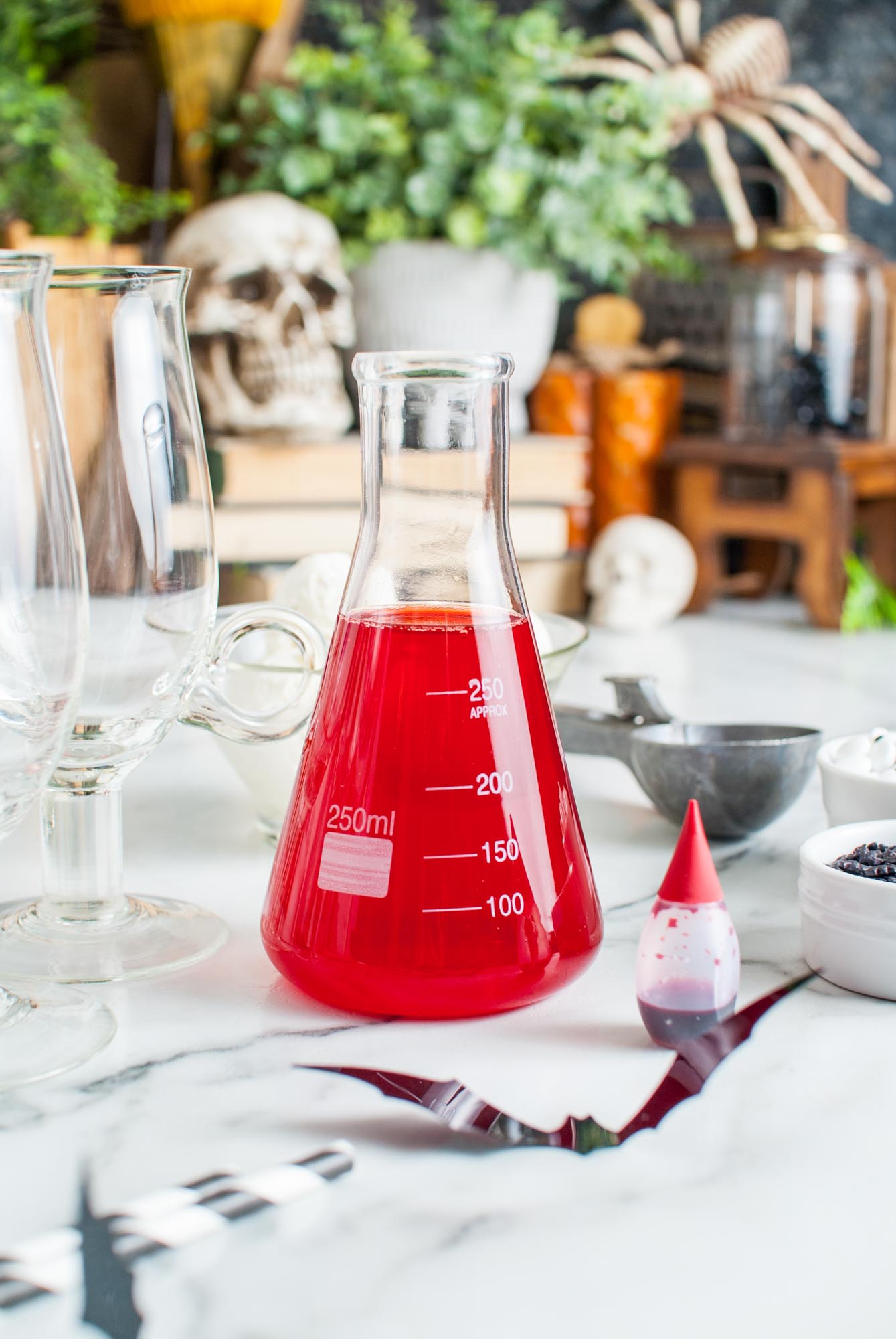 a glass erlenmeyer flask filled with fruit punch soda on a table with a bottle of red food coloring. Spooky decor is in the background.