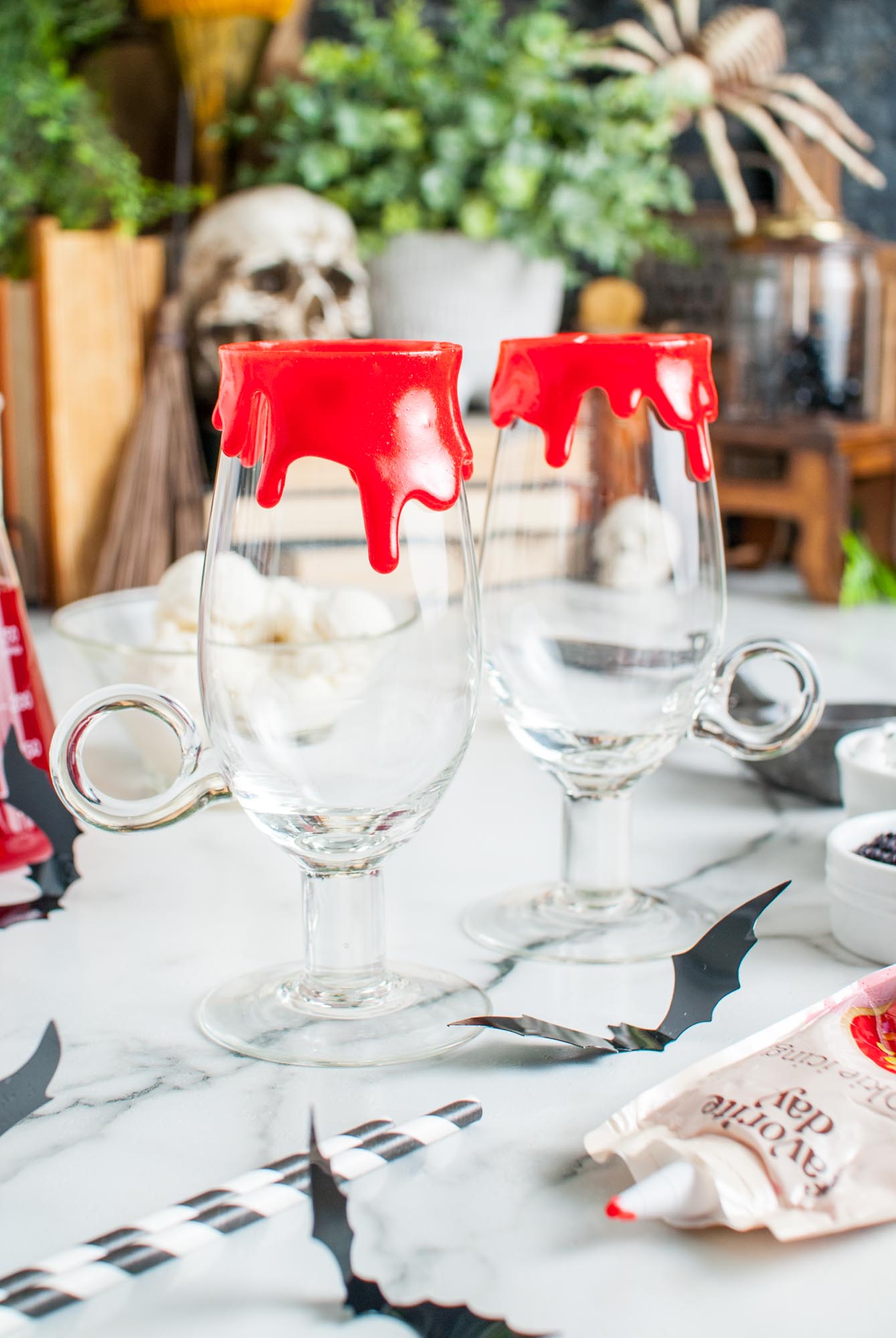 Two footed glass mugs rimmed with red icing to look like dripping blood. Halloween decorations are in the background.