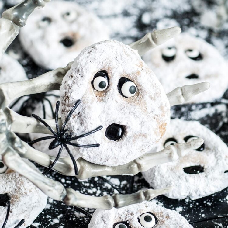 ghost-faced peanut butter cookies decorated with powdered sugar, black icing, and candy eyes. One cookie is held up by a skeleton hand.