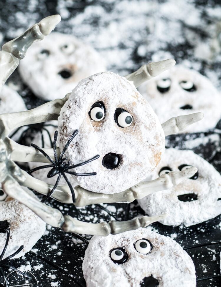 ghost-faced peanut butter cookies decorated with powdered sugar, black icing, and candy eyes. One cookie is held up by a skeleton hand.