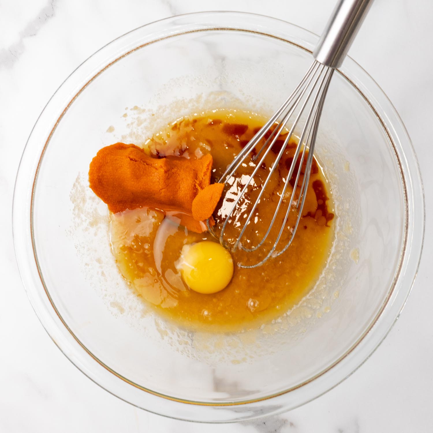 a glass bowl holding melted butter, sugar, pumpkin puree, an egg, and a whisk.