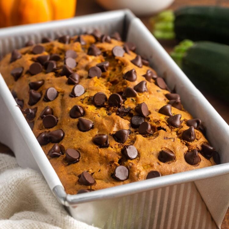 a pan of pumpkin bread with chocolate chips. In the background is a small pumpkin and fresh zucchinis.