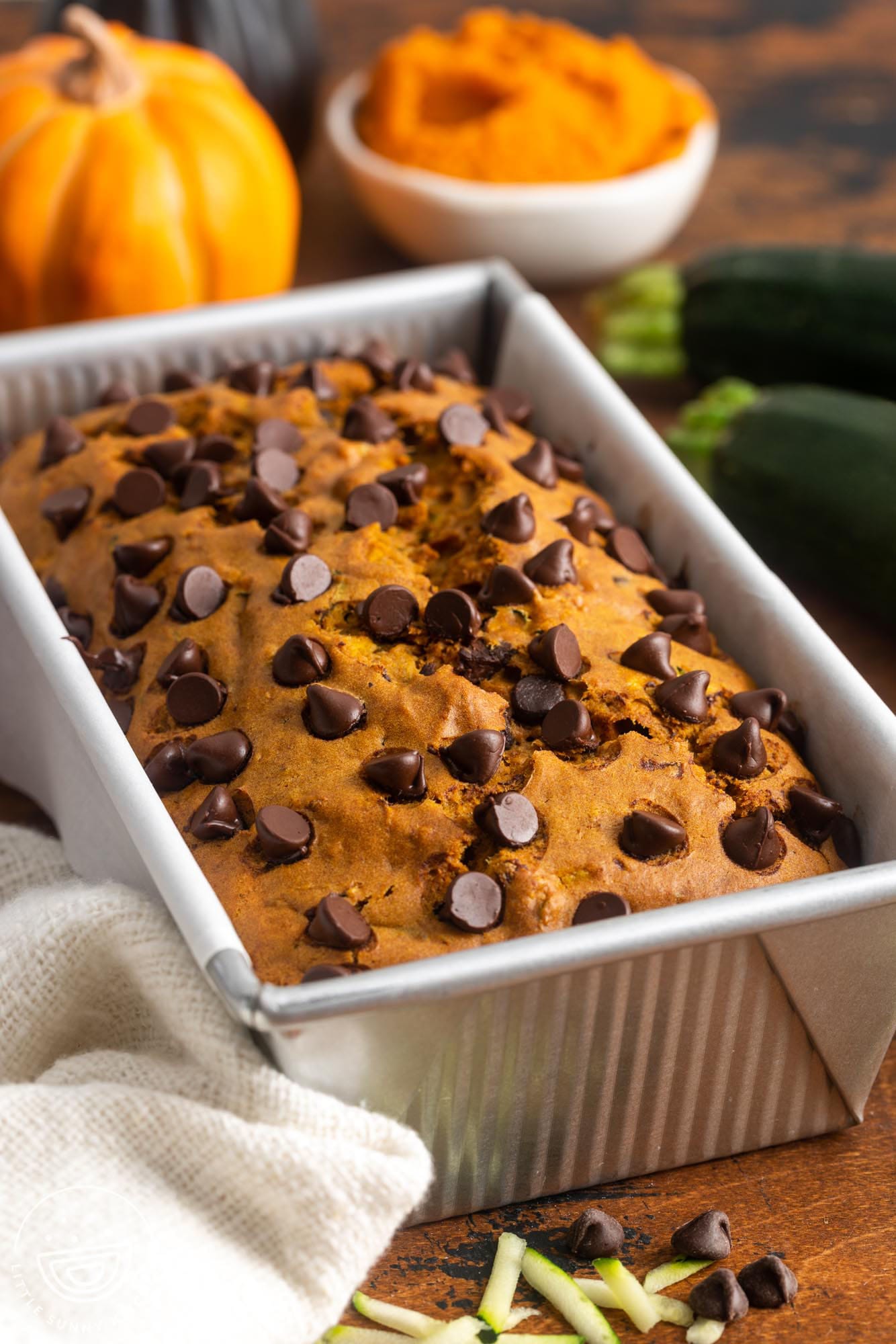 a pan of pumpkin bread with chocolate chips. In the background is a small pumpkin and fresh zucchinis.