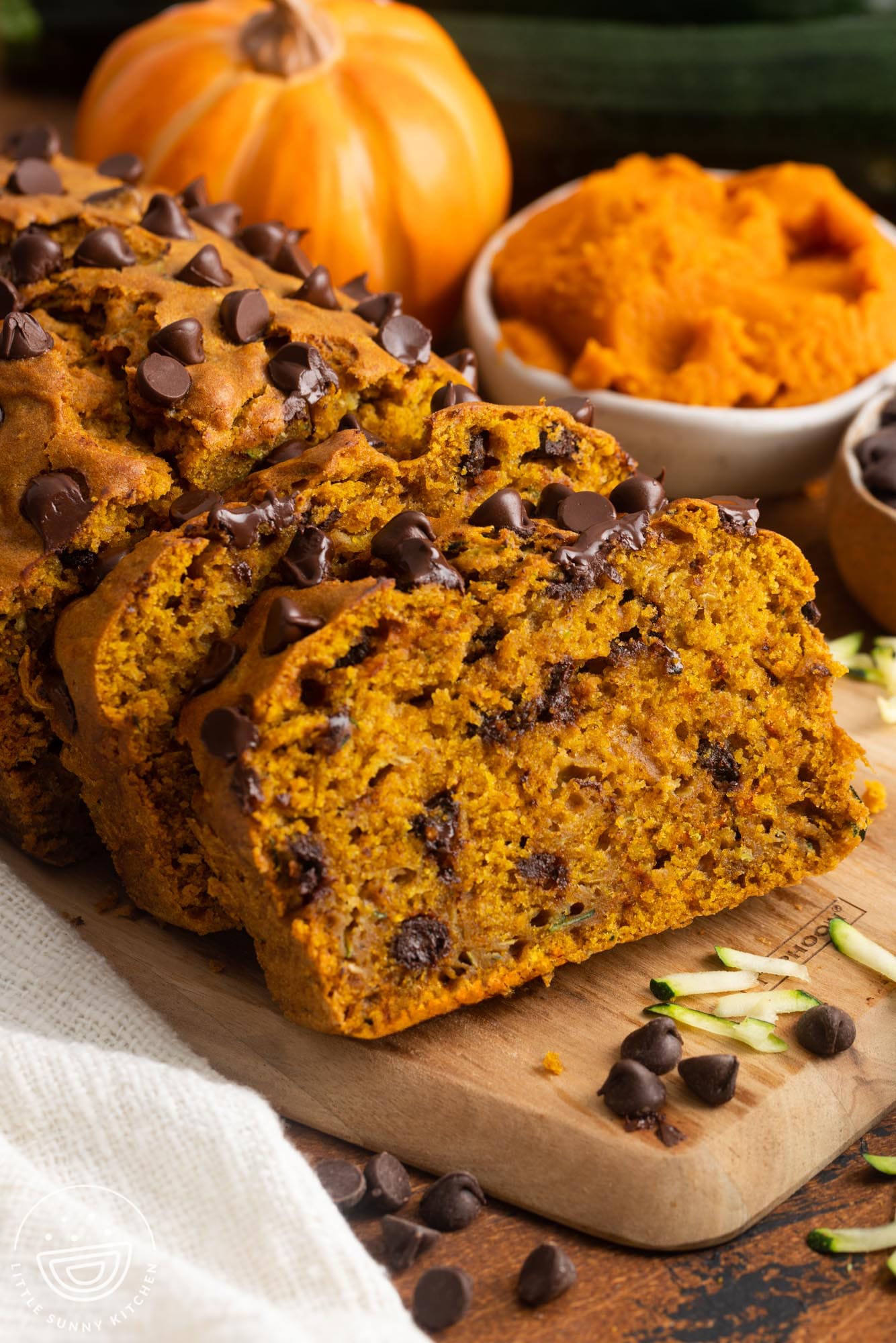 slices of pumpkin bread with zucchini and chocolate chips on a wooden cutting board. In the background is a small pumpkin and pumpkin puree in a bowl.