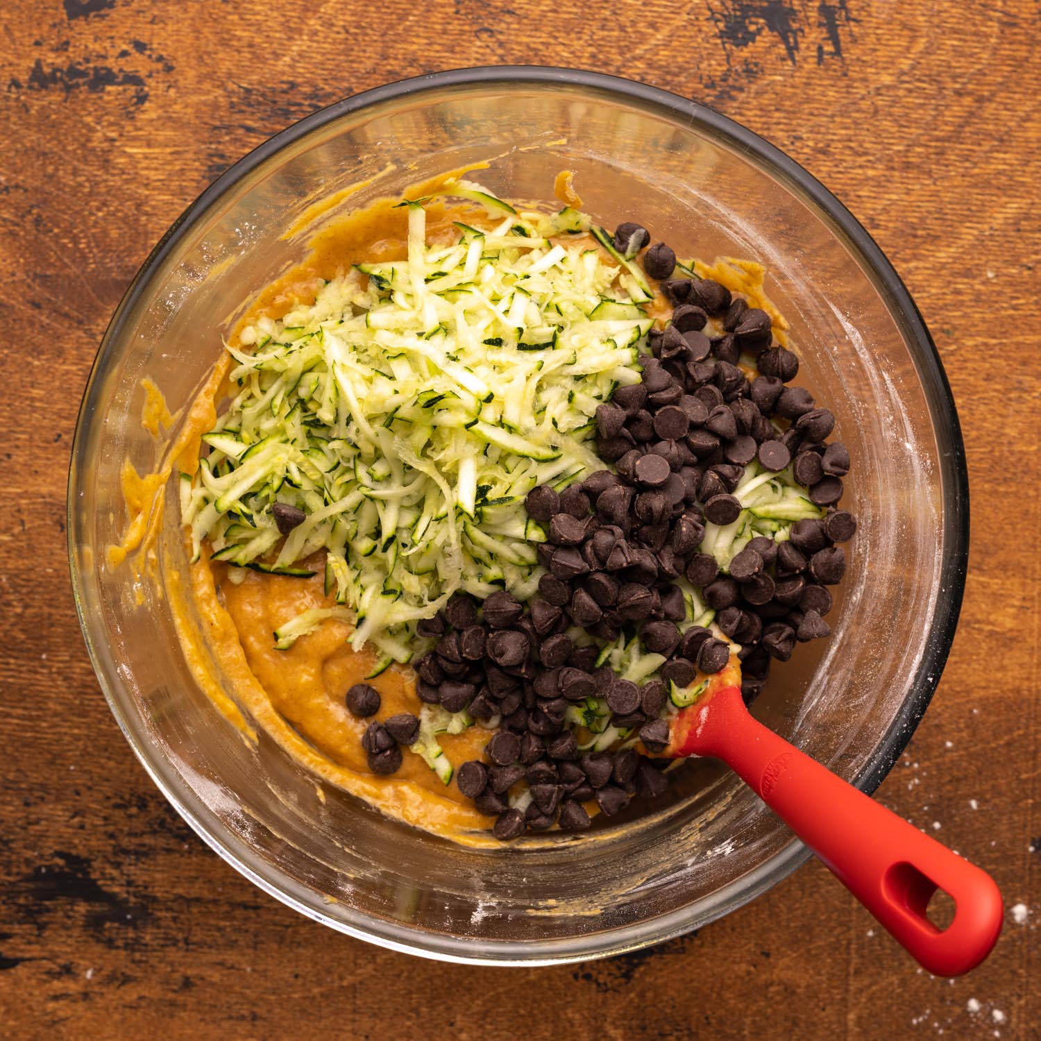 shredded zucchini and chocolate chips added to pumpkin bread batter in a glass bowl with a red spoon.