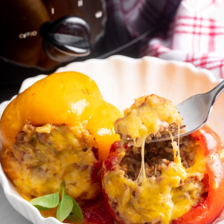 one yellow and one red stuffed pepper in a white bowl. A fork is eating the filling from the red one.