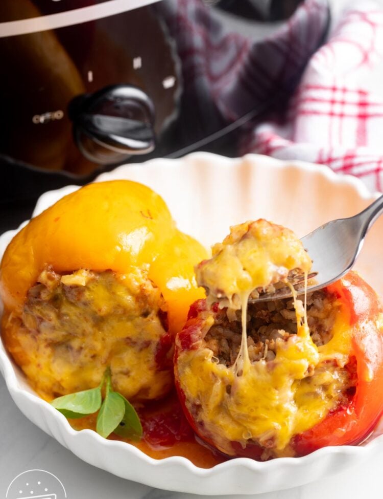 one yellow and one red stuffed pepper in a white bowl. A fork is eating the filling from the red one.