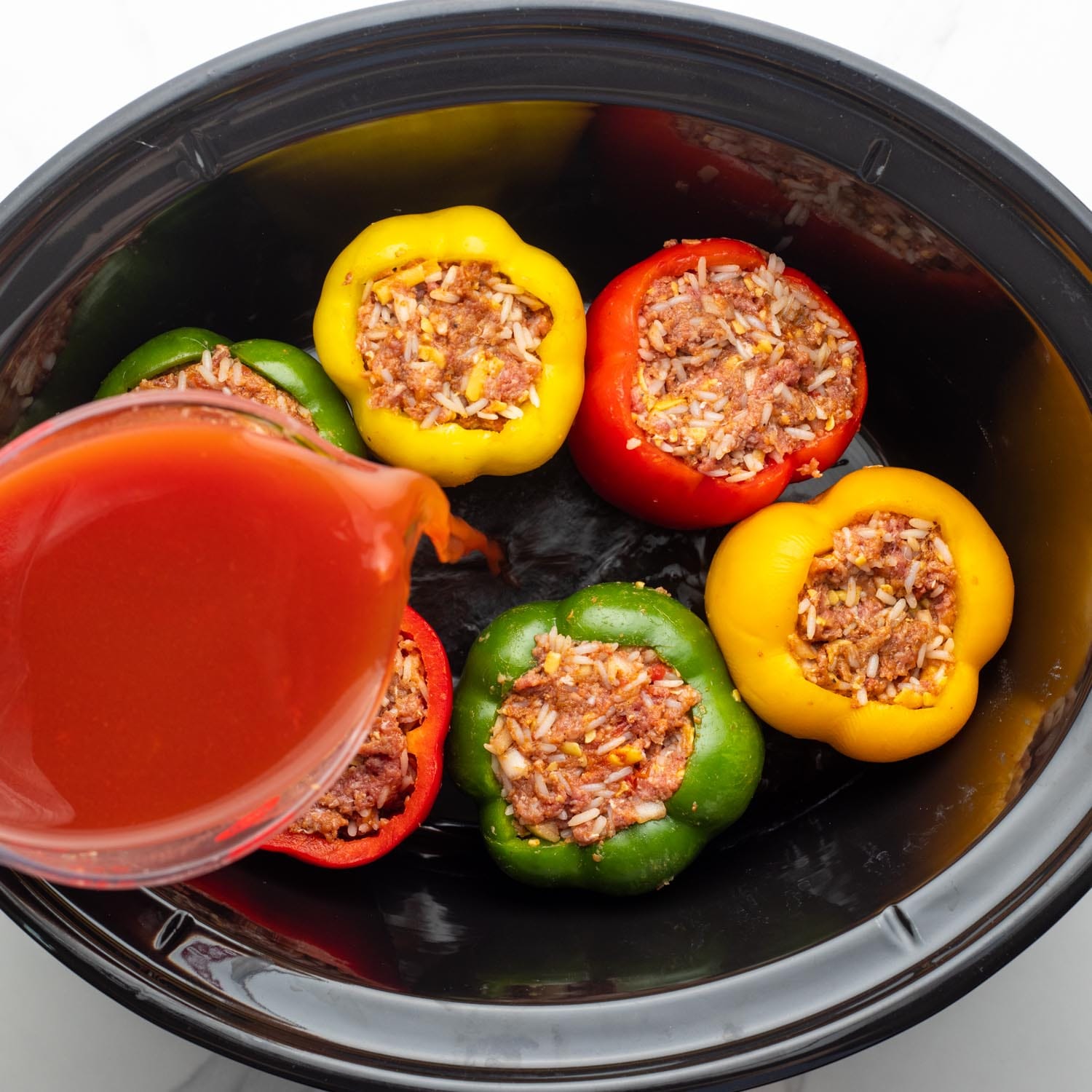 Stuffed bell peppers in a large slow cooker. Tomato juice is being poured in.