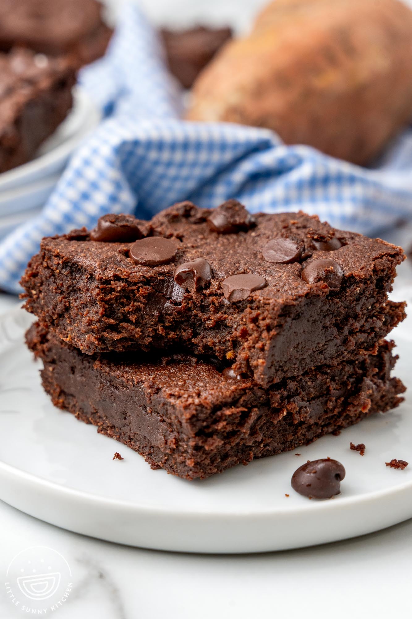 two dense and chewy sweet potato brownies with chocolate chips stacked on a white plate. The top brownie has a bite taken.