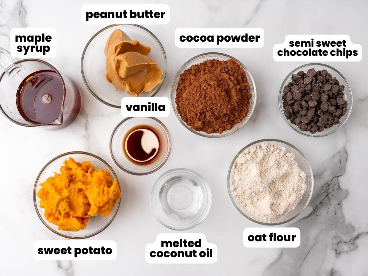 Ingredients for sweet potato brownies in small glass bowls on a marble counter. From top left: maple syrup, peanut butter, cocoa powder, semisweet chocolate chips, oat flour, melted coconut oil, sweet potato, and vanilla extract.