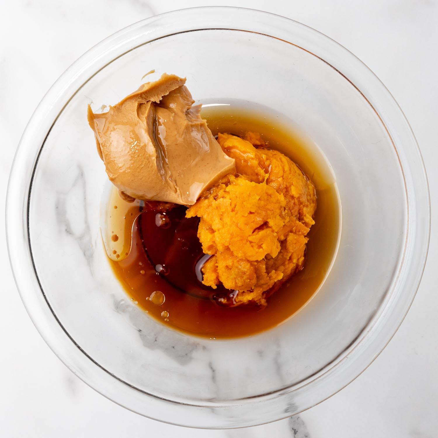 mashed sweet potato, peanut butter, maple syrup, and melted coconut oil in a glass mixing bowl, viewed from above.