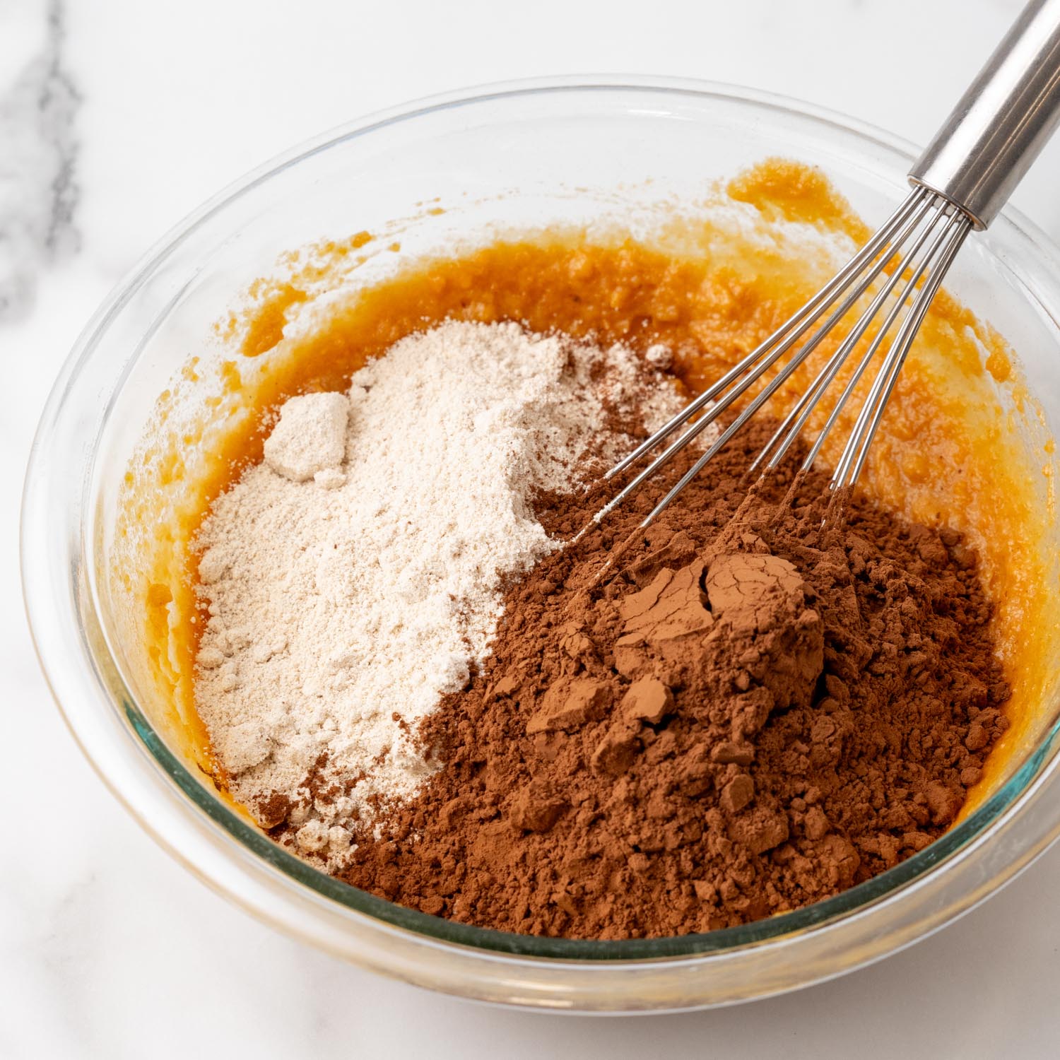 flour and cocoa powder added to sweet potato puree mixture in a glass mixing bowl with a whisk.