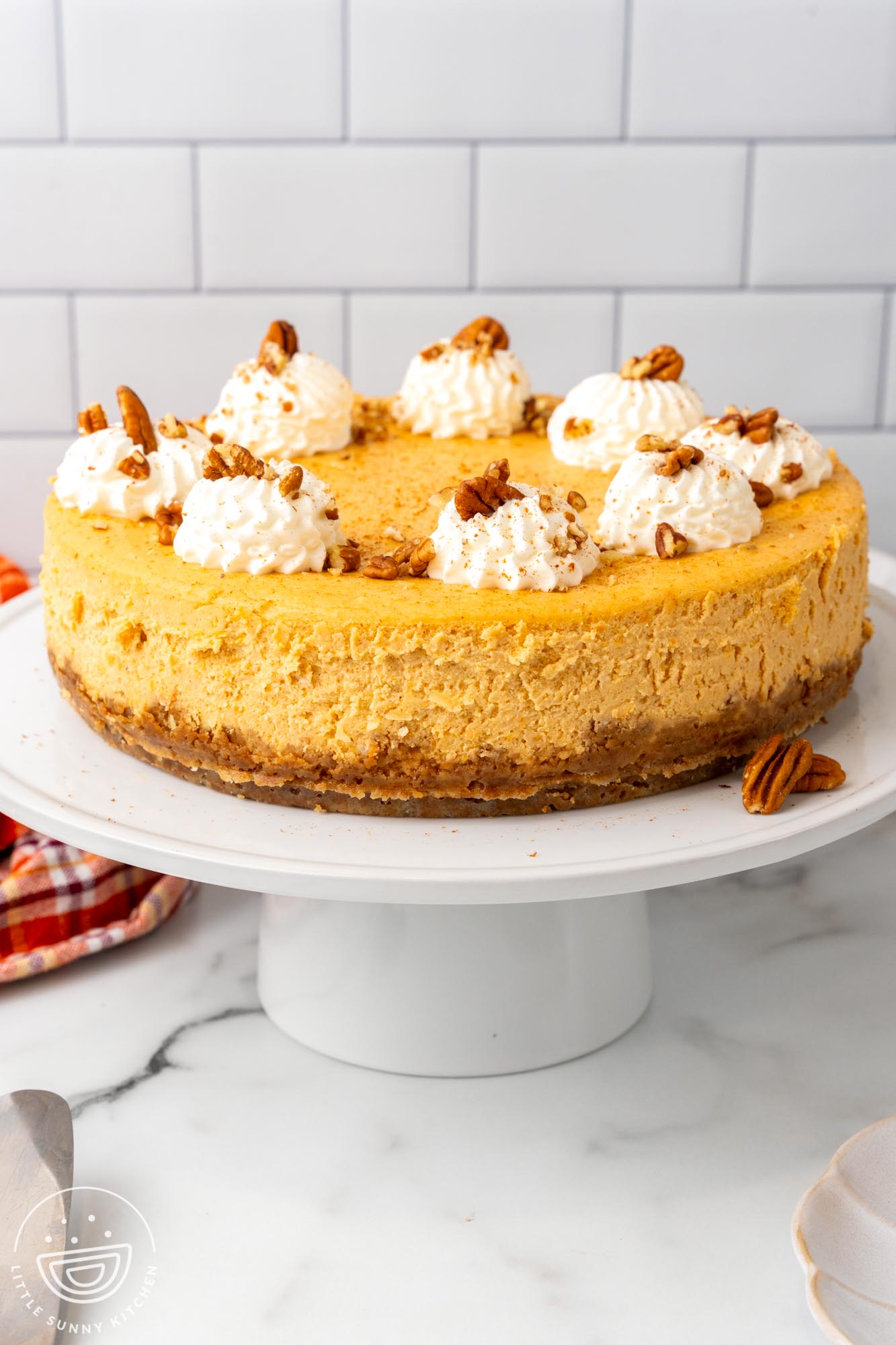 a whole sweet potato cheesecake topped with dollops of whipped cream and chopped pecans. The cheesecake has a thick crust made with gingersnaps and is staged on a cake pedestal.