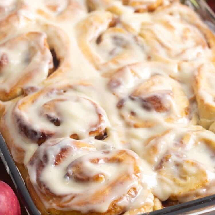 apple pie cinnamon rolls in a glass baking dish topped with icing. Around the pan are fresh green and red apples.