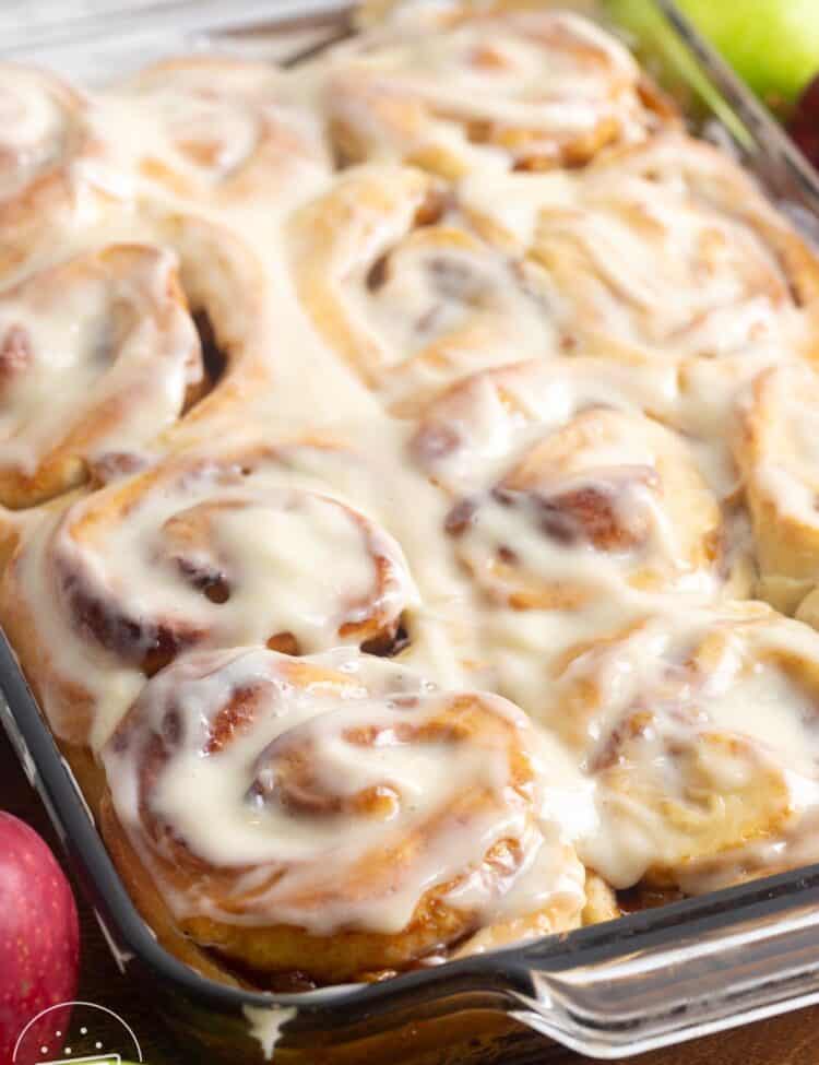 apple pie cinnamon rolls in a glass baking dish topped with icing. Around the pan are fresh green and red apples.