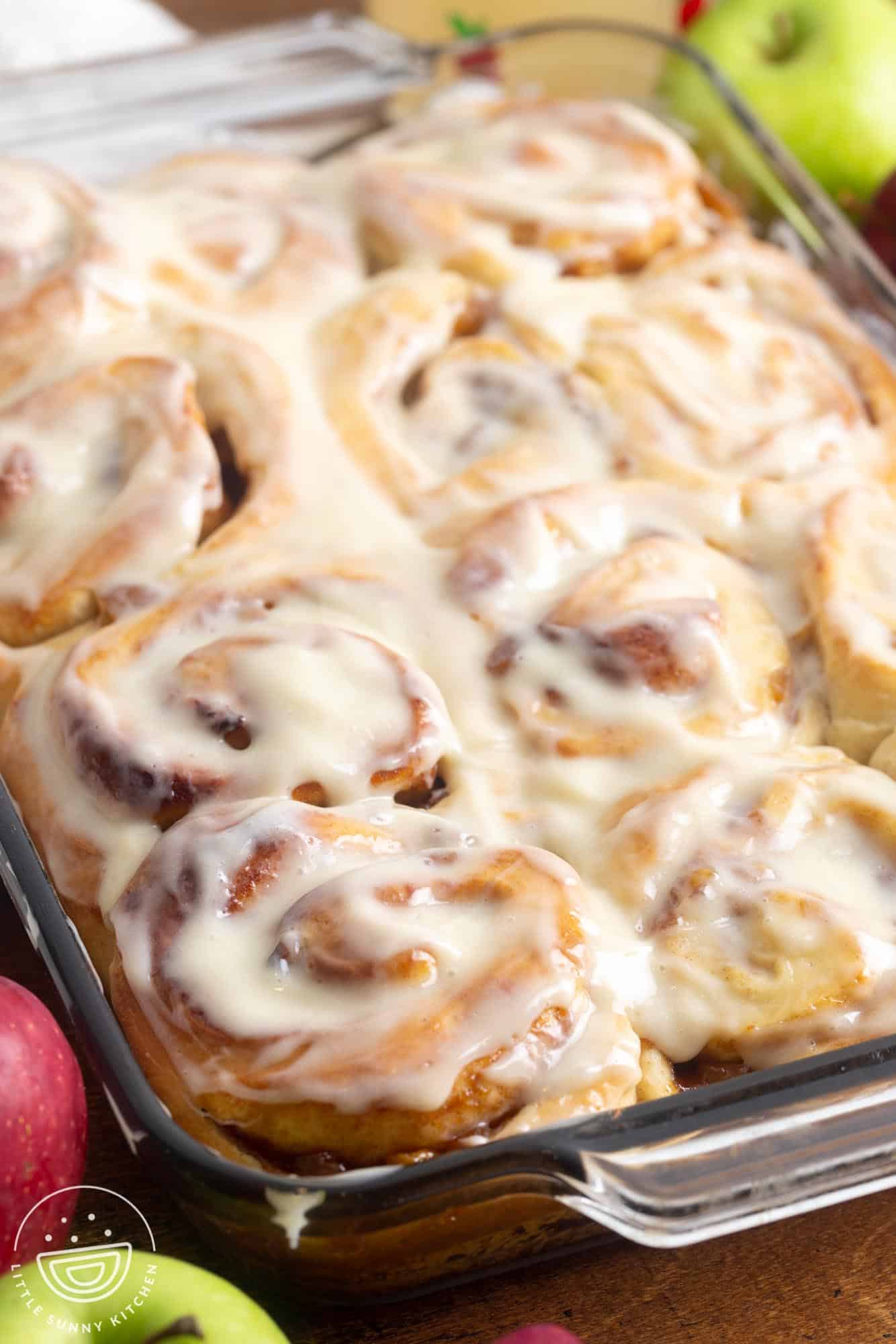 apple pie cinnamon rolls in a glass baking dish topped with icing. Around the pan are fresh green and red apples.