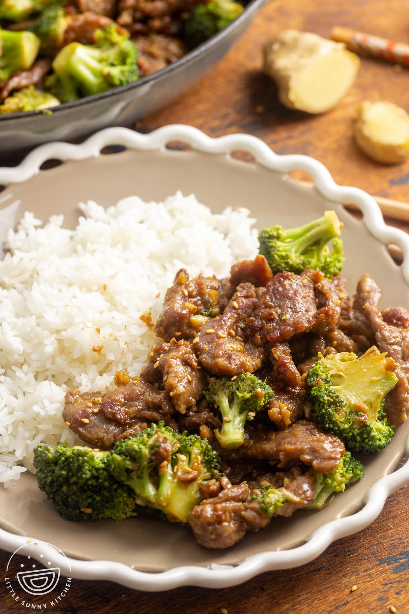 a white shallow bowl with scalloped edges holding a serving of rice and a serving of beef and broccoli stir fry topped with sesame seeds.