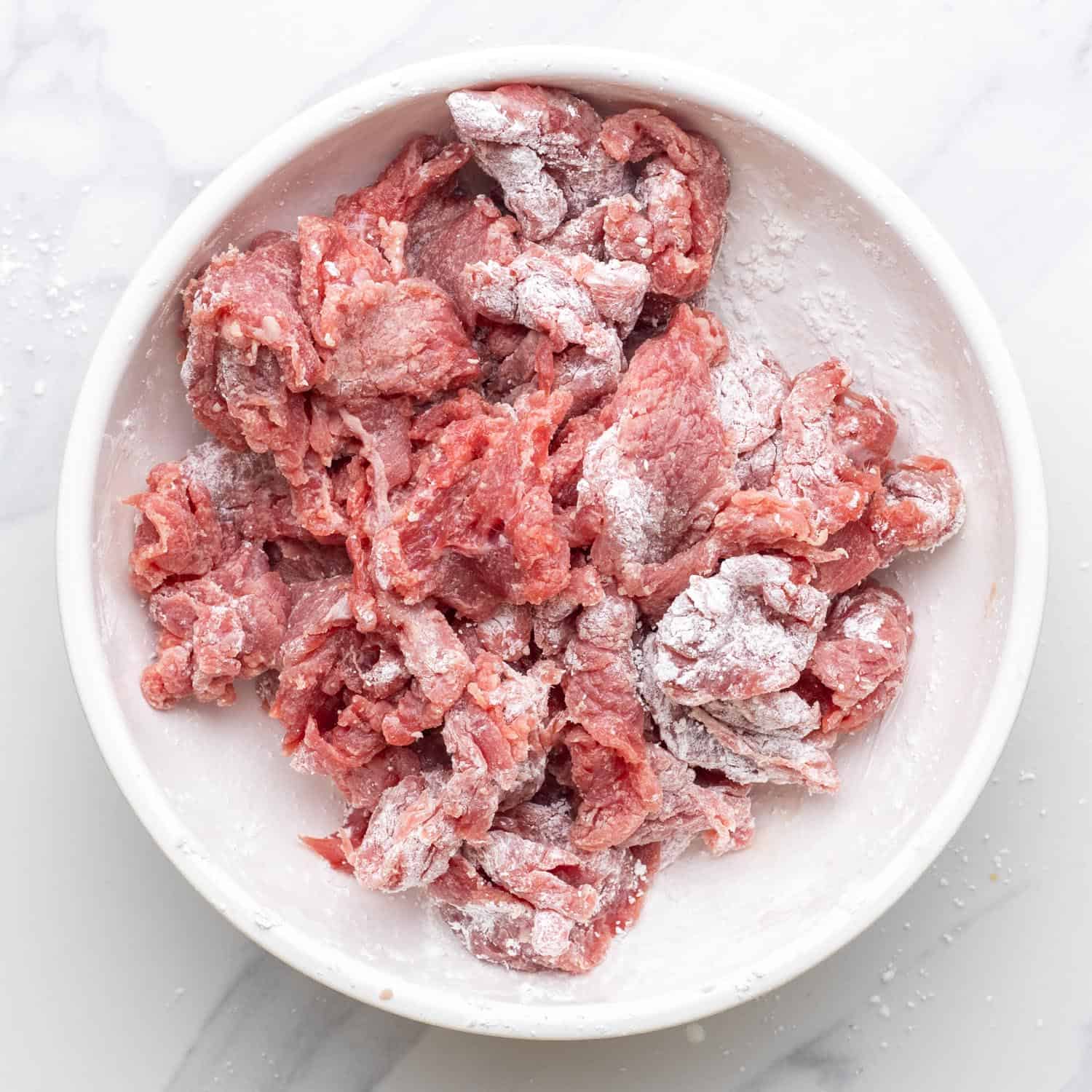 beef strips dredged with corn starch in a white bowl.