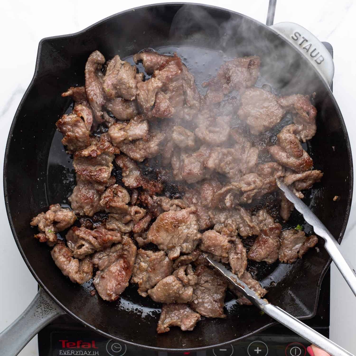 beef strips stir fried in a cast iron skillet with tongs.