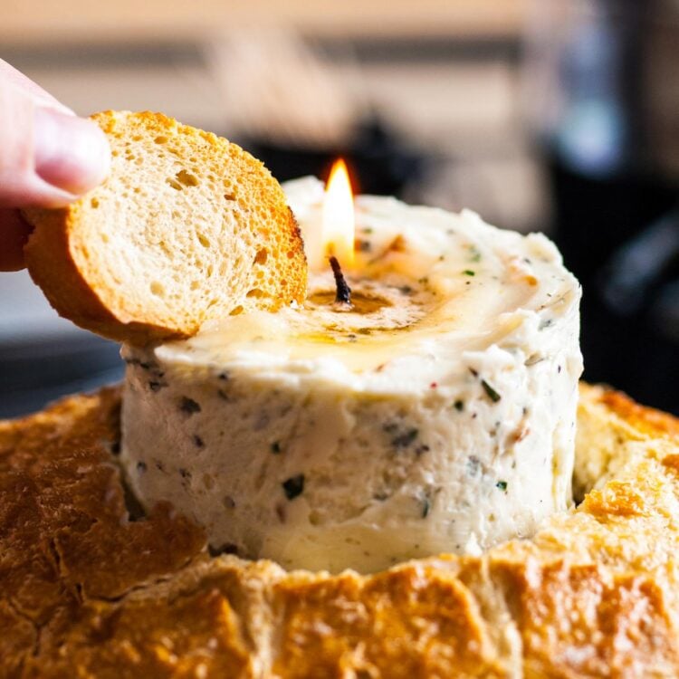 a hand holding a piece of bread into a lit butter candle that is standing up inside of a loaf of crusty bread.