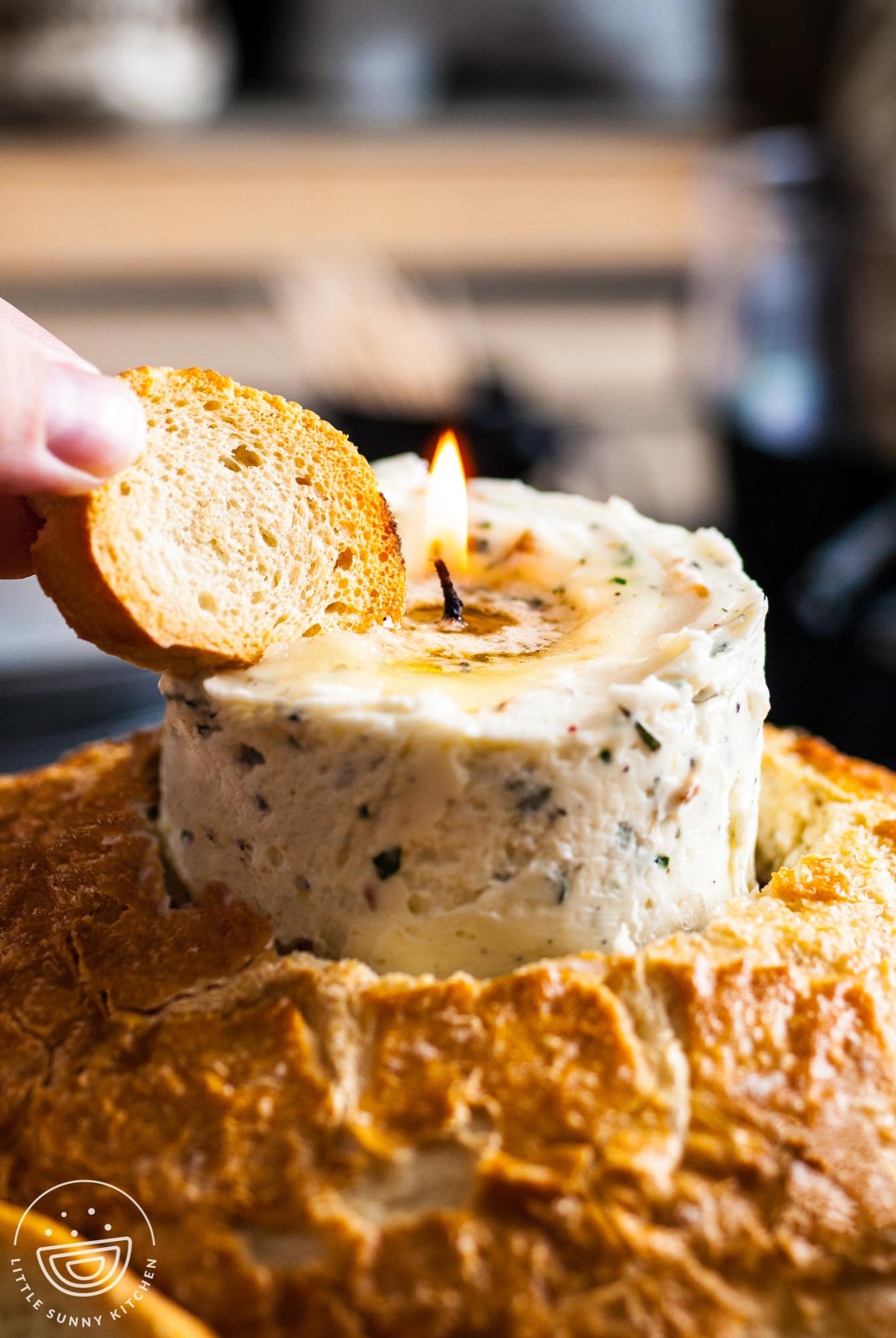 a hand holding a piece of bread into a lit butter candle that is standing up inside of a loaf of crusty bread.