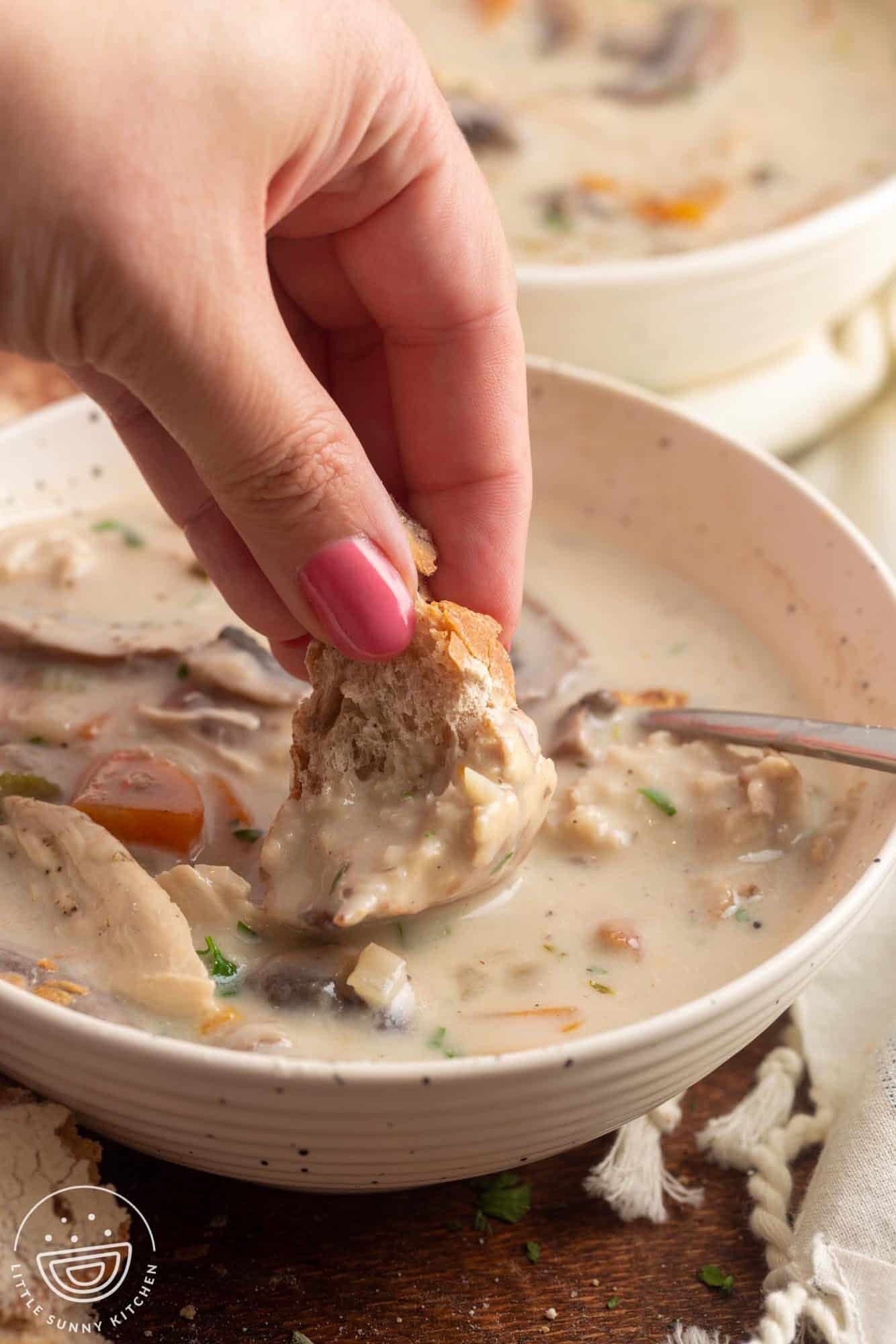 a hand dipping a piece of bread into a bowl of creamy chicken soup.