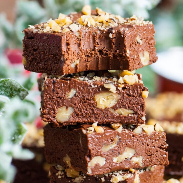 four squares of chocolate fantasy fudge with chopped walnuts, stacked on top of each other. The top piece has a bite taken. In the background is holiday greenery.