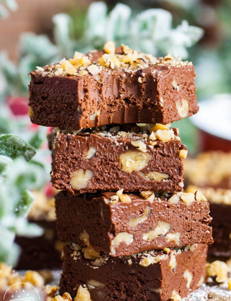 four squares of chocolate fantasy fudge with chopped walnuts, stacked on top of each other. The top piece has a bite taken. In the background is holiday greenery.