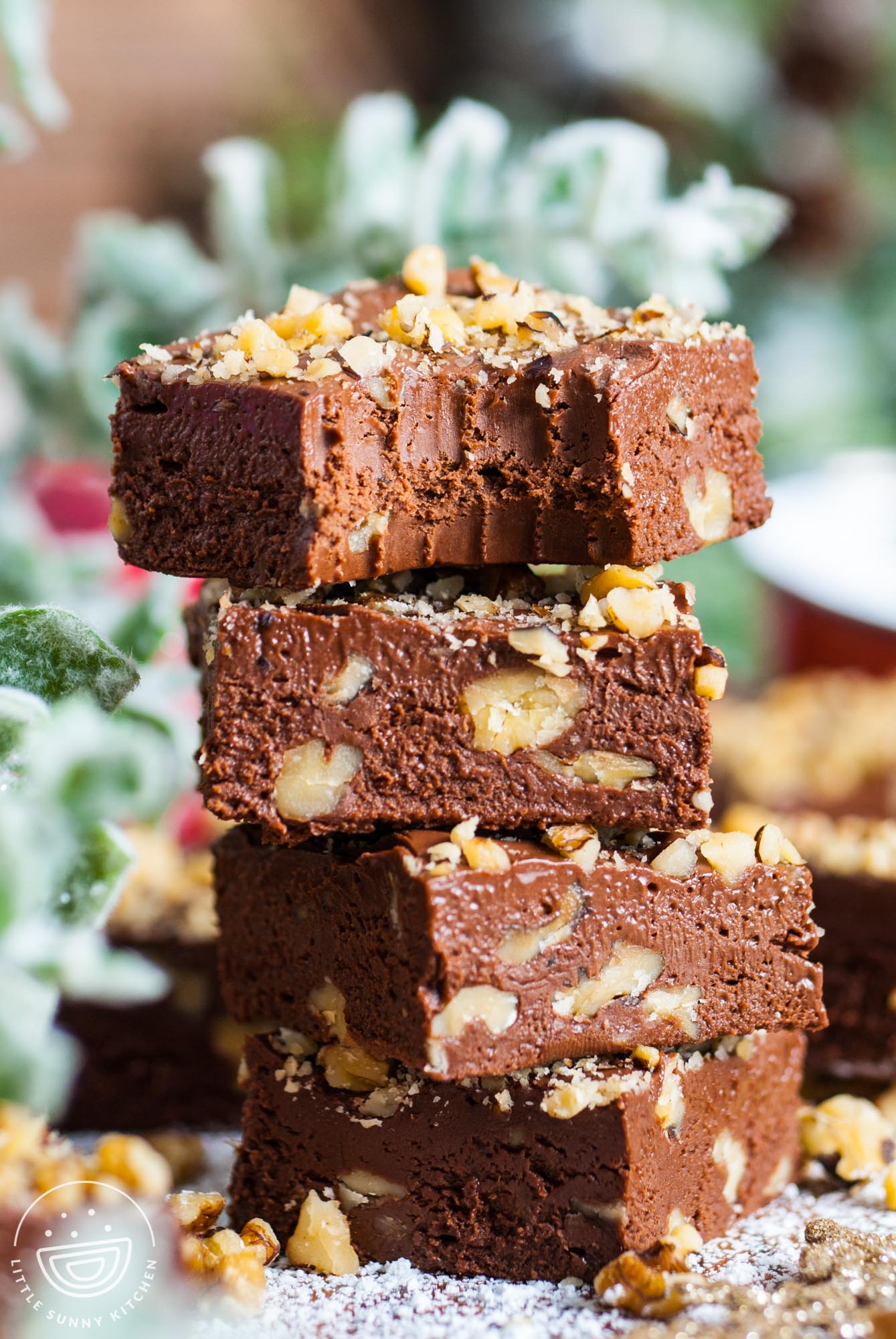 four squares of chocolate fantasy fudge with chopped walnuts, stacked on top of each other. The top piece has a bite taken. In the background is holiday greenery.