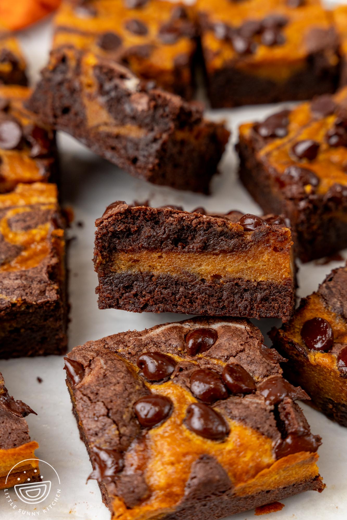 swirled pumpkin brownies with melty chocolate chips, cut into squares on a piece of parchment paper. Two brownies are tipped to show the layering inside.