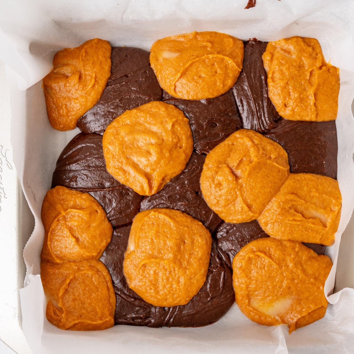 a pan lined with parchment paper, holding brownie batter and pumpkin mixture.