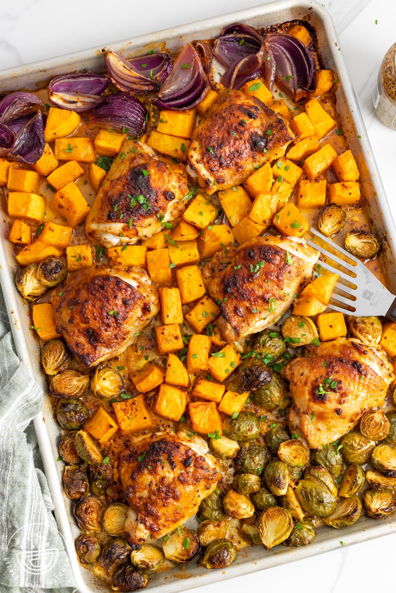 overhead view of a sheet pan filled with baked chicken thighs, red onion, butternut squash cubes, and brussel sprouts, all in a honey mustard marinade.