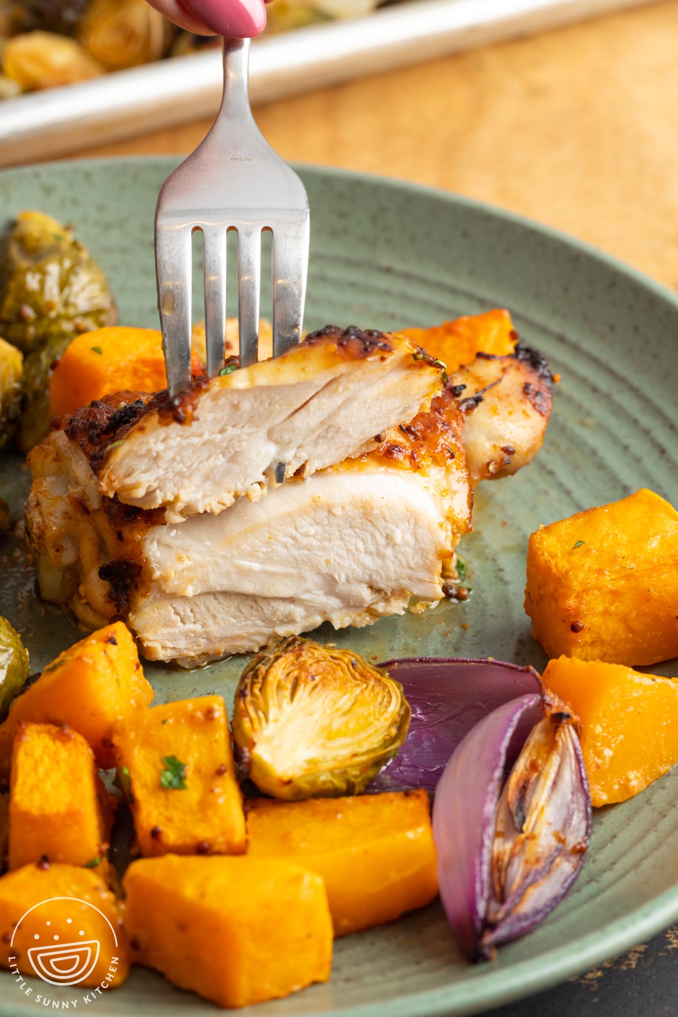 a fork holding a piece of chicken thigh to show how perfectly cooked it is. On the side are roasted brussel sprouts and squash.