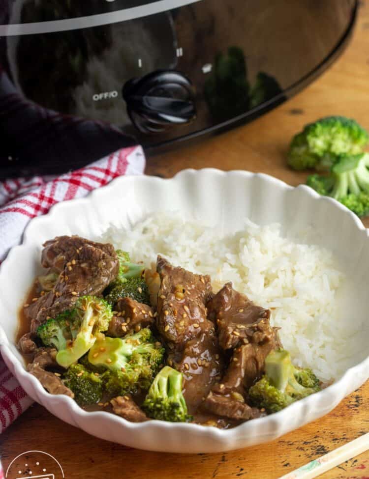 a bowl of rice with beef and broccoli, topped with sesame seeds. The bowl is nest to a black crock pot.