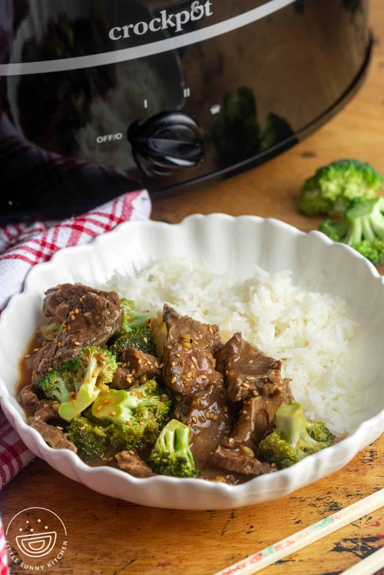 a bowl of rice with beef and broccoli, topped with sesame seeds. The bowl is nest to a black crock pot.