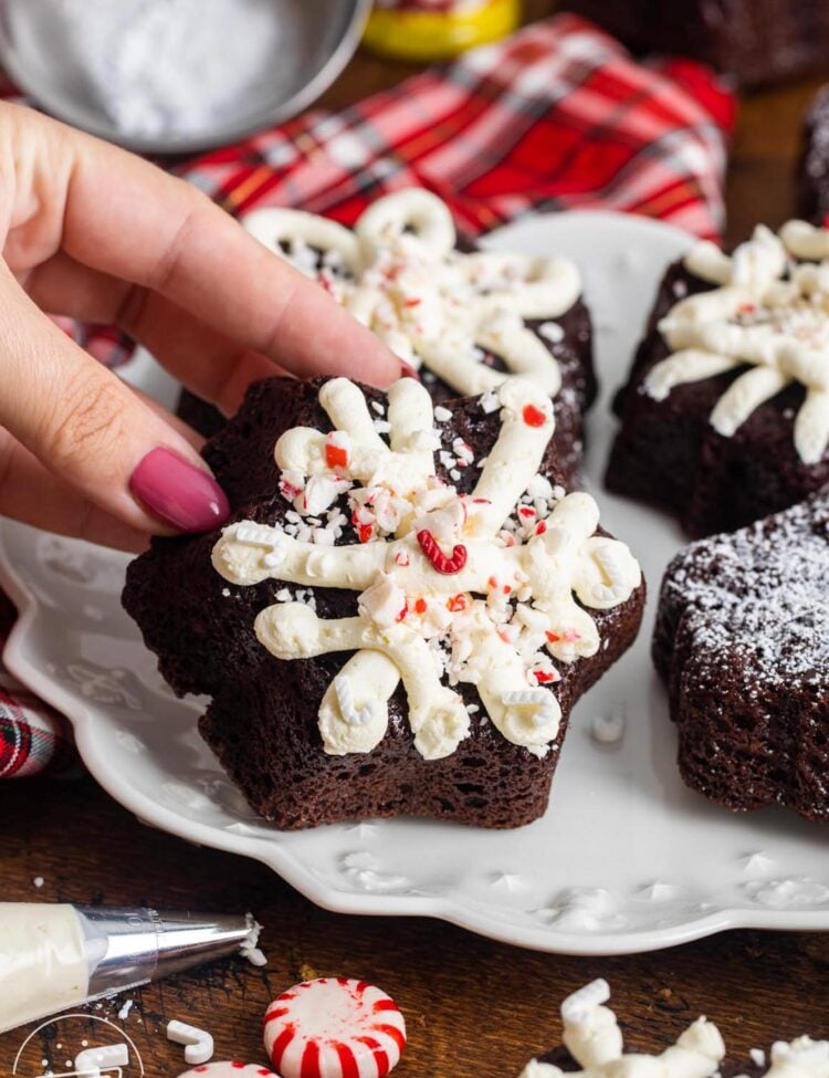 crushed peppermints and holiday sprinkles on top of chocolate mini cakes with white frosting. A hand is holding one up.