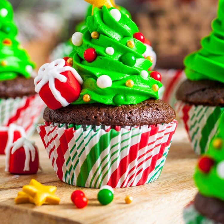 chocolate cupcakes in paper liners. Each has a swirl of icing in the shape of a tree on top and a gold star. sprinkles are added to look like ornaments and fondant packages are on the side.