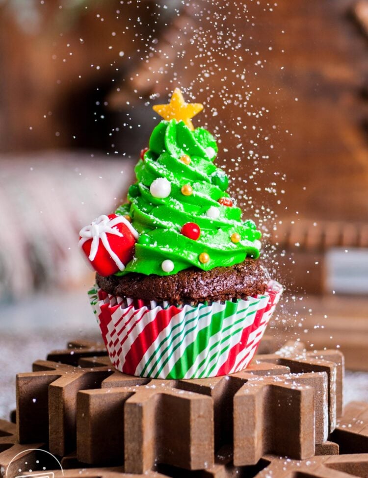 a christmas tree cupcake on top of a wooden snowflake. Powdered sugar is sprinkled over to look like snow.